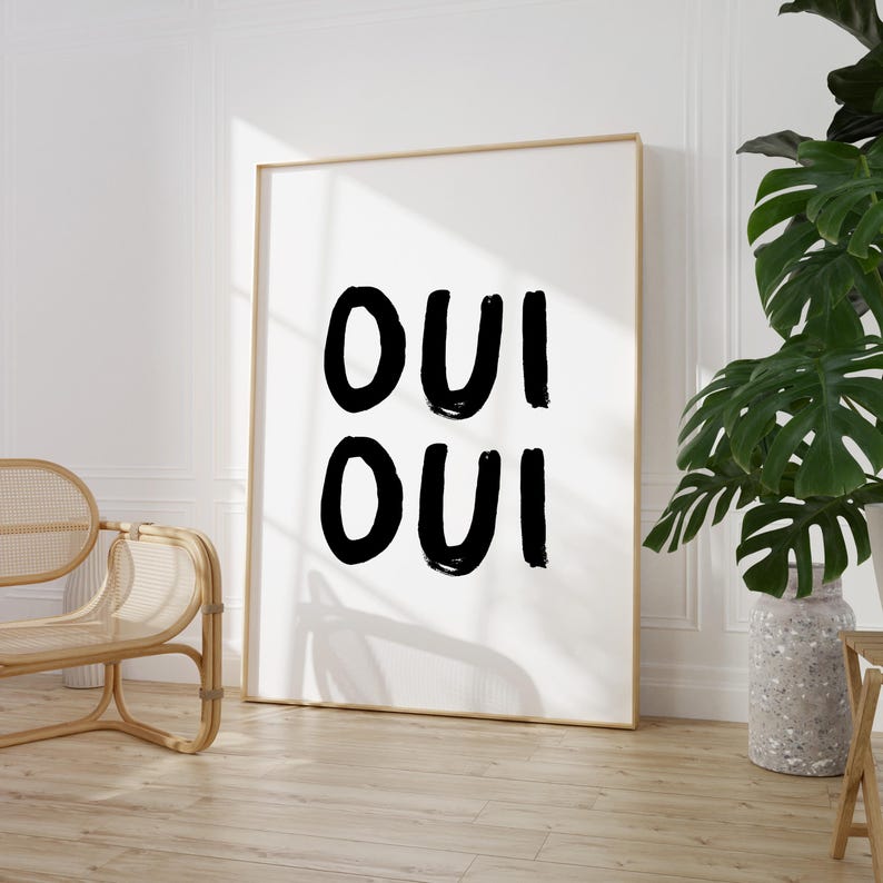 May include: A black and white art print with the word "OUI" written twice in a bold, brushstroke font. The print is framed in a thin, gold frame and is hanging on a white wall.