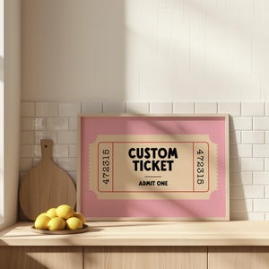 May include: A framed pink print featuring a custom ticket design. The ticket is tan with red border details and the words "CUSTOM TICKET" and "ADMIT ONE" in black. A wooden cutting board and a plate of lemons are in the foreground.