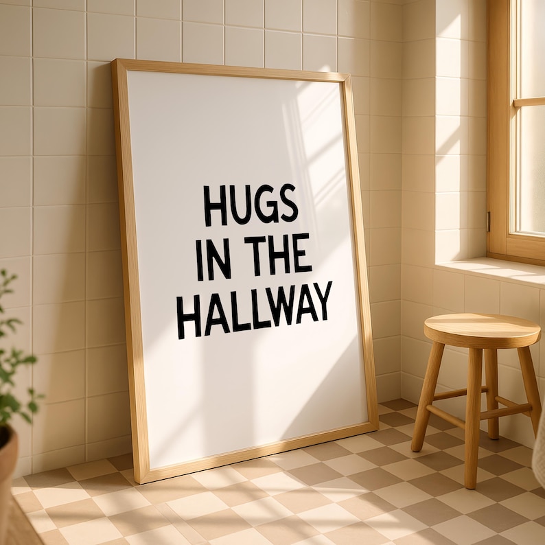 Op de afbeelding: Een ingelijste print met de tekst "HUGS IN THE HALLWAY" in zwarte letters. De lijst is van licht hout en de print leunt tegen een betegelde muur. Een kleine houten kruk staat in de buurt.