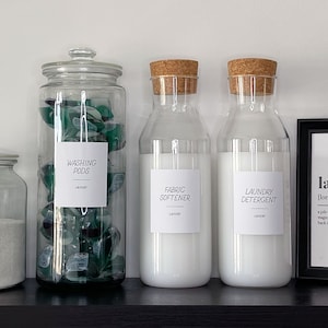 May include: Four glass jars with labels for laundry supplies: Oxi Powder, Washing Pods, Fabric Softener, and Laundry Detergent. The jars are on a black shelf with a framed print that reads "laundry [lom-dree] noun"
