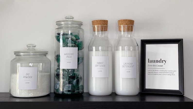 May include: Four glass jars with labels for laundry supplies: Oxi Powder, Washing Pods, Fabric Softener, and Laundry Detergent. The jars are on a black shelf with a framed print that reads "laundry [lom-dree] noun"