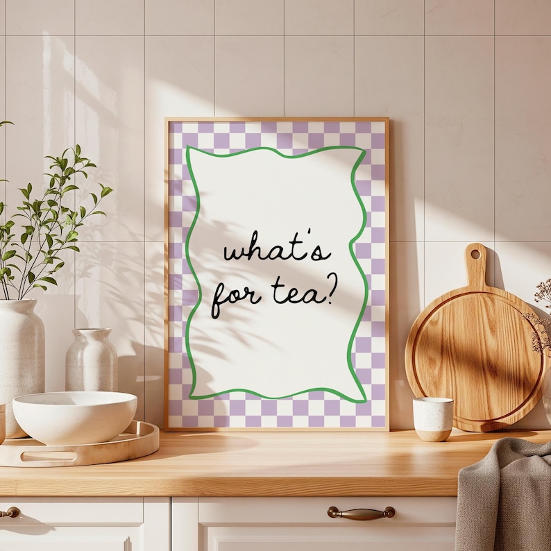 Peut inclure: Une affiche encadr&eacute;e avec le texte "what's for tea?" en &eacute;criture noire. L'affiche a un centre blanc avec une bordure verte ondul&eacute;e et un fond &agrave; carreaux violets et blancs. L'affiche est expos&eacute;e sur un comptoir en bois.