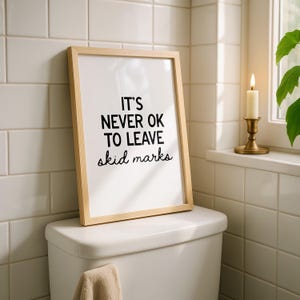 May include: A framed print with the text "IT'S NEVER OK TO LEAVE skid marks" in black on a white background. The frame is light wood. The print is displayed on a white toilet in a bathroom setting.