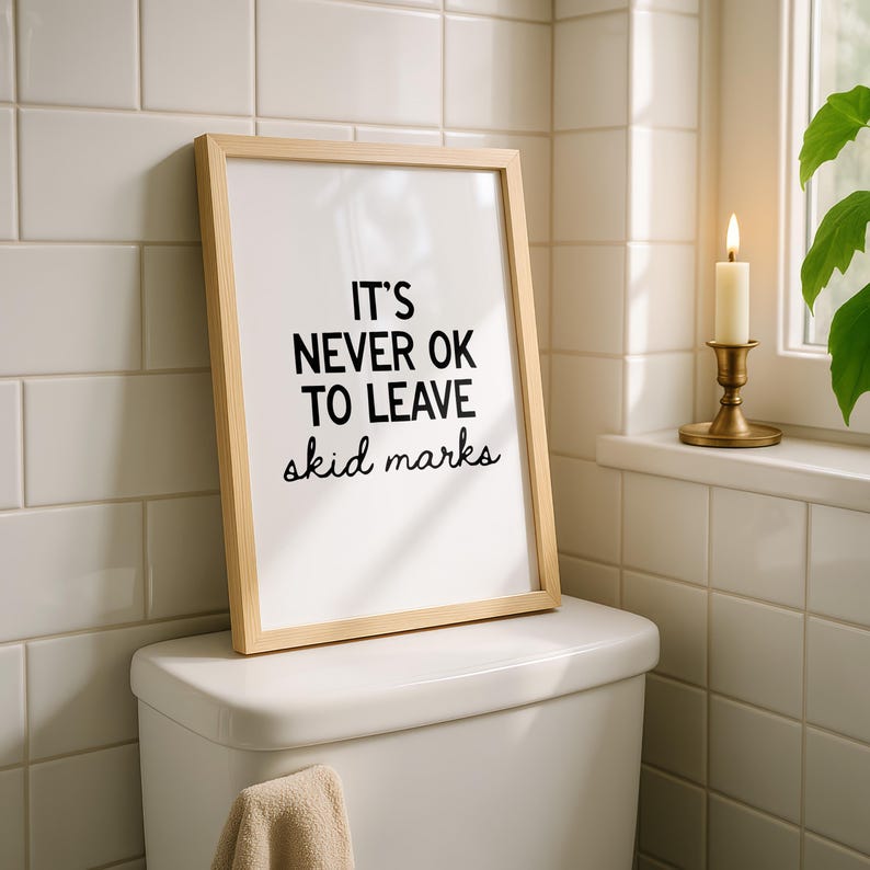 May include: A framed print with the text "IT'S NEVER OK TO LEAVE skid marks" in black on a white background. The frame is light wood. The print is displayed on a white toilet in a bathroom setting.