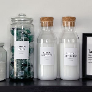 May include: Four glass jars with labels on a black shelf. The jars contain laundry detergent, fabric softener, washing pods, and oxi powder. A framed print with the word "laundry" and its definition is on the shelf.