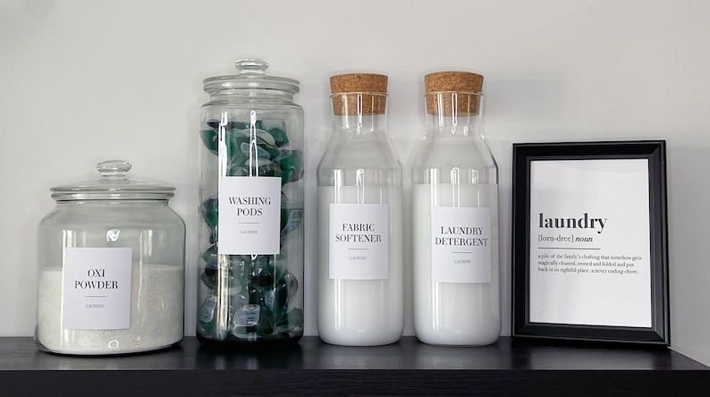 May include: Four glass jars with labels on a black shelf. The jars contain laundry detergent, fabric softener, washing pods, and oxi powder. A framed print with the word "laundry" and its definition is on the shelf.