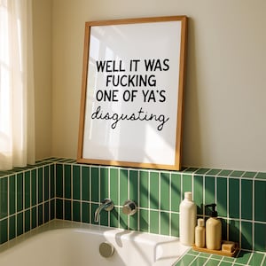 May include: A framed print with the text "WELL IT WAS FUCKING ONE OF YA'S disgusting" in black lettering. The print is in a light wood frame and is displayed above a bathtub with green tile.