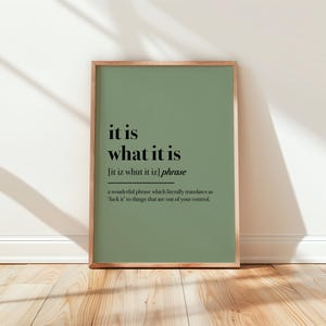 May include: A framed art print with a sage green background and the phrase "it is what it is" in black text. The print includes a phonetic pronunciation and a definition. The frame is a light brown color.