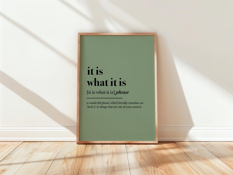 May include: A framed art print with a sage green background and the phrase "it is what it is" in black text. The print includes a phonetic pronunciation and a definition. The frame is a light brown color.
