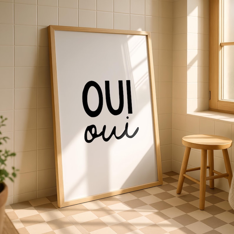 May include: A framed art print with the word "OUI" in large black letters above "oui" in a smaller, cursive font. The artwork has a light wood frame and is leaning against a tiled wall. A small wooden stool sits nearby.