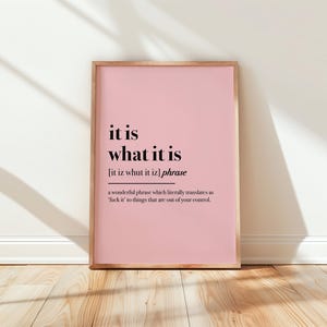 May include: A framed pink print with the phrase "it is what it is" in black text. The print includes a definition and sits on a wooden floor against a white wall.