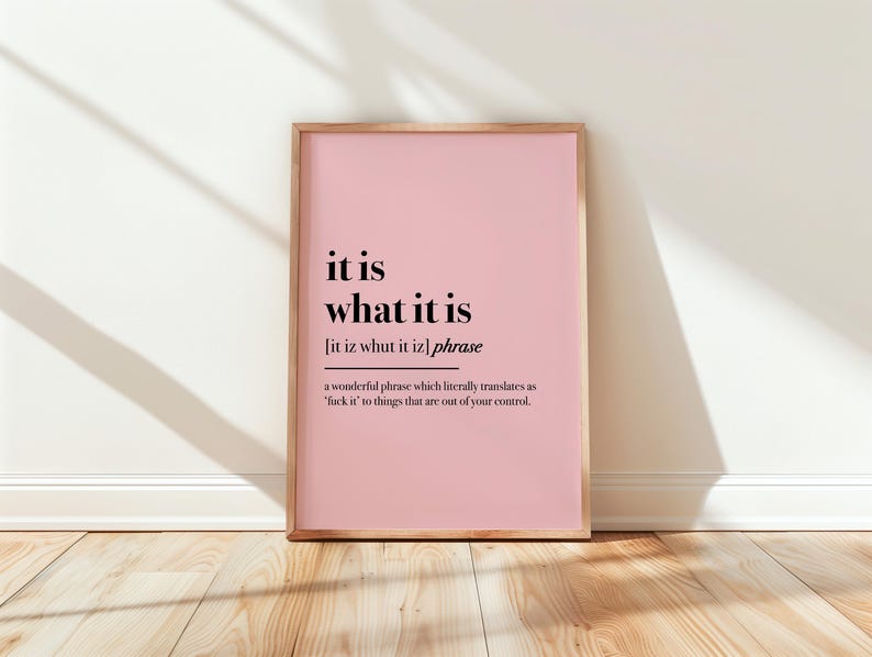 May include: A framed pink print with the phrase "it is what it is" in black text. The print includes a definition and sits on a wooden floor against a white wall.