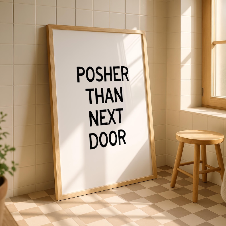 Op de afbeelding: Een ingelijste print met de zwarte tekst "POSHER THAN NEXT DOOR" leunt tegen een betegelde muur. De lijst is van licht hout en de kamer heeft een neutraal kleurenpalet met een kleine houten kruk.