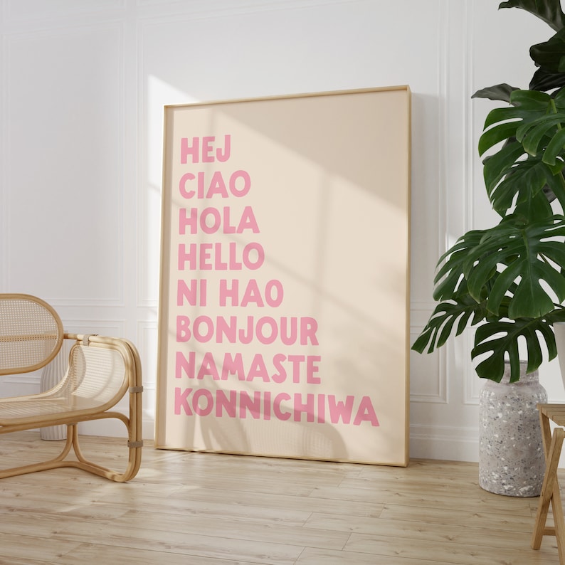 Peut inclure: Une affiche beige avec un cadre dor&eacute; pr&eacute;sentant les mots "Hej", "Ciao", "Hola", "Hello", "Ni Hao", "Bonjour", "Namaste" et "Konnichiwa" en lettres roses.