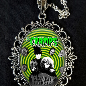 May include: A silver necklace with a round pendant featuring a green and yellow spiral design. The pendant has a black and white image of a band called "The Cramps" with the text "Gravest Tribute".