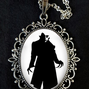 May include: A silver necklace with a cameo pendant featuring a black silhouette of a vampire with sharp claws. The cameo is set in a silver filigree frame.