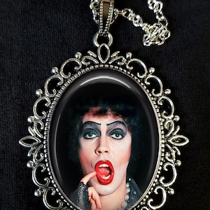 May include: A silver necklace with a cameo pendant featuring a portrait of Dr. Frank-N-Furter from the movie The Rocky Horror Picture Show. The cameo is set in a silver filigree frame.