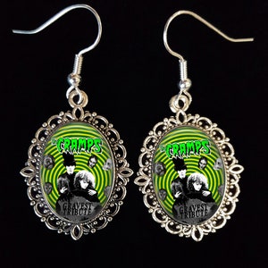 May include: A pair of silver earrings with oval-shaped green and black cameo pendants featuring a band with the text "The Cramps" and a graphic of a band with the text "Gravest Tribute".