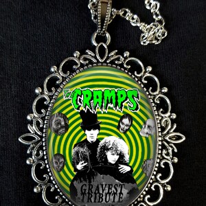 May include: A silver necklace with a round pendant featuring a green and black image of The Cramps band. The image includes the band's name, a swirling green and black background, and the text "Gravest Tribute".