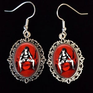 May include: A pair of silver earrings with oval-shaped red glass cabochons featuring a black and white illustration of a woman sitting on a drum set with the word "Cramps" printed on the drum.