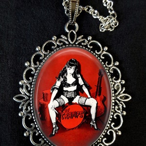 May include: A silver pendant necklace with a red oval cameo featuring a woman in a black and white lingerie outfit sitting on a drum with the word "The Cramps" printed on it.