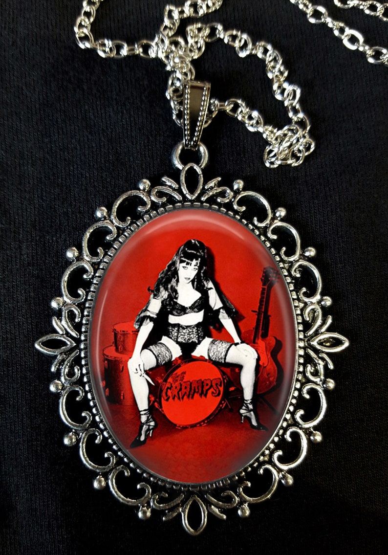 May include: A silver pendant necklace with a red oval cameo featuring a woman in a black and white lingerie outfit sitting on a drum with the word "The Cramps" printed on it.