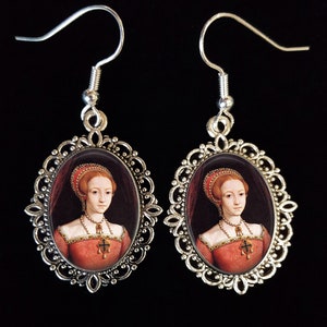 May include: Silver earrings with oval-shaped portraits of a woman in a red dress and a white head covering. The portraits are set in a silver filigree frame.