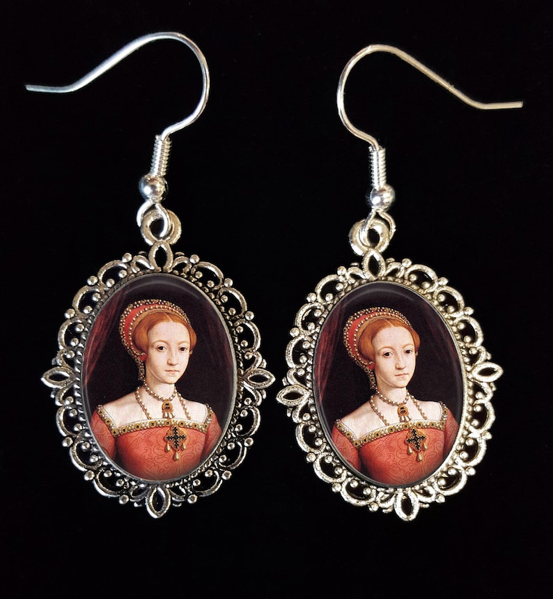May include: Silver earrings with oval-shaped portraits of a woman in a red dress and a white head covering. The portraits are set in a silver filigree frame.