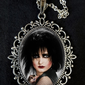 May include: A silver oval pendant necklace with a black and white image of a woman with dark hair and dark eye makeup. The pendant is surrounded by a decorative silver frame.