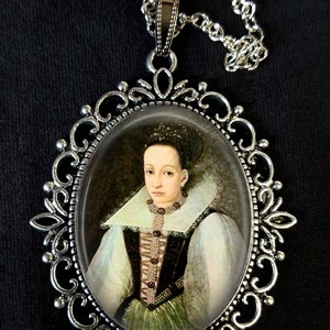 Op de afbeelding: Een zilveren ketting met een ovale hanger met een portret van een vrouw in een witte kanten kraag en een groene jurk.