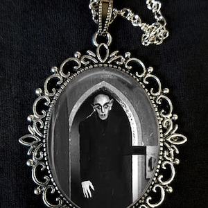 Puede incluir: Un collar de plata con un colgante de camafeo que presenta una imagen en blanco y negro de un hombre con un traje oscuro de pie en una puerta. El camafeo está engastado en un marco de plata adornado.