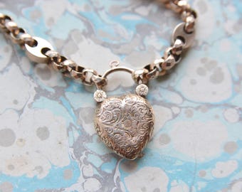 May include: A gold chain necklace with a heart-shaped locket. The locket is engraved with a floral design.