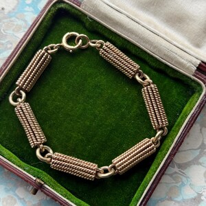 May include: A gold-toned bracelet with a series of cylindrical, rope-like segments connected by small links. The bracelet is displayed inside a vintage, brown jewelry box with a green velvet lining.