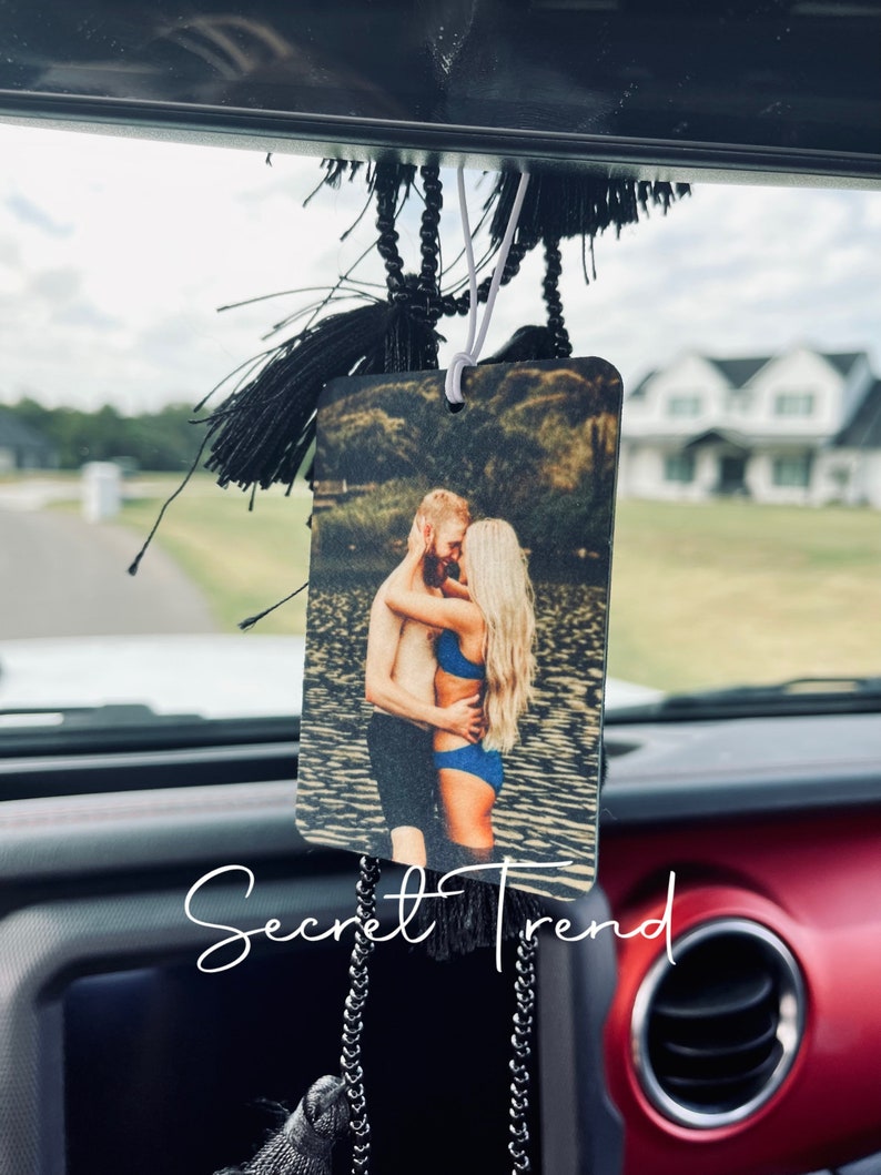 May include: A car air freshener with a photo of a couple hugging on a beach. The photo is printed on a rectangular piece of wood with rounded corners. The air freshener is hanging from a black tassel.