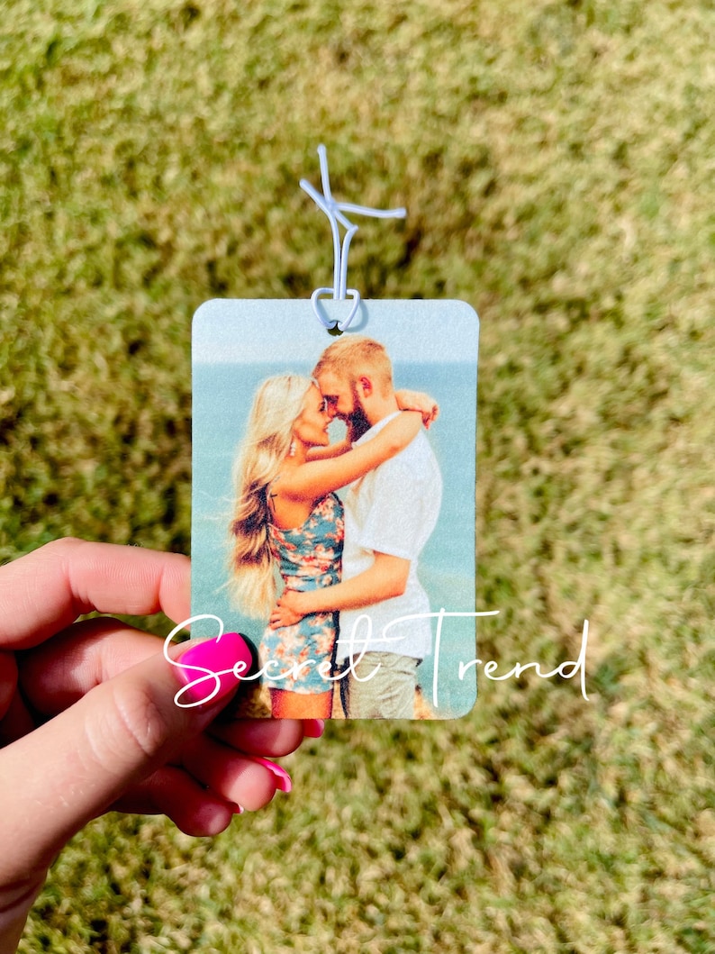 May include: A white car air freshener with a photo of a couple hugging on a beach. The text "Secret Trend" is printed on the bottom of the air freshener.