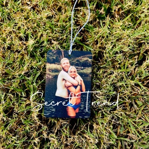 May include: A photo car air freshener with a couple hugging in front of a lake. The couple is wearing swimsuits and the photo is printed on a black background.