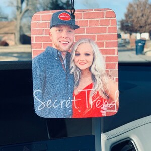 May include: A car air freshener with a photo of a couple in front of a brick wall. The man is wearing a black baseball cap with a red logo that says "Hooey". The woman is wearing a red dress. The text "Secret Trend" is written below the photo.