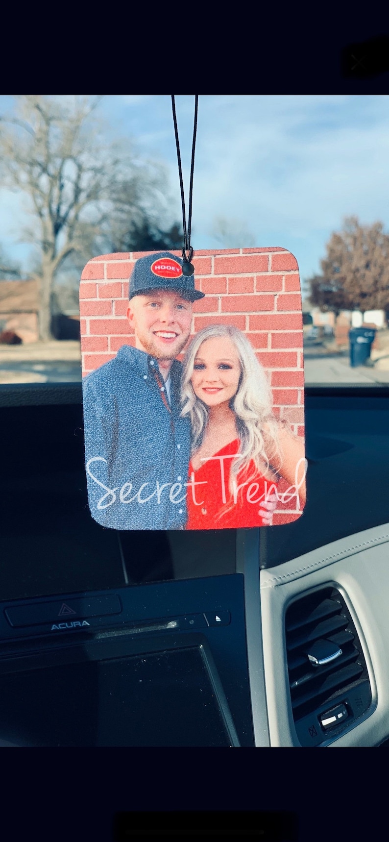 May include: A car air freshener with a photo of a couple in front of a brick wall. The man is wearing a black baseball cap with a red logo that says "Hooey". The woman is wearing a red dress. The text "Secret Trend" is written below the photo.