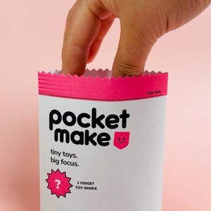 May include: A white and pink "pocket make" package with the text "tiny toys. big focus." and "1 fidget toy inside." A hand reaches into the package. The package has a pink top and bottom.