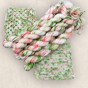 May include: A collection of green, white and pink yarn skeins. The yarn is twisted and piled together. There are three crocheted squares in the image, one is white with green and pink accents, one is green with white and pink accents, and one is white with green and pink accents.