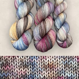 May include: Three skeins of variegated yarn featuring a blend of blue, brown, pink, and cream tones. Below the skeins is a swatch of knitted fabric displaying the same multi-coloured pattern in a consistent stockinette stitch texture.