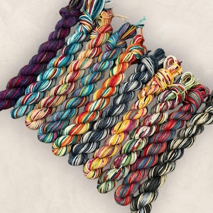 May include: A collection of ten twisted skeins of yarn arranged in a diagonal row. The skeins feature a variety of color combinations including solid deep purple, teal and white, orange and yellow, dark grey, and multi-colored variegated patterns.