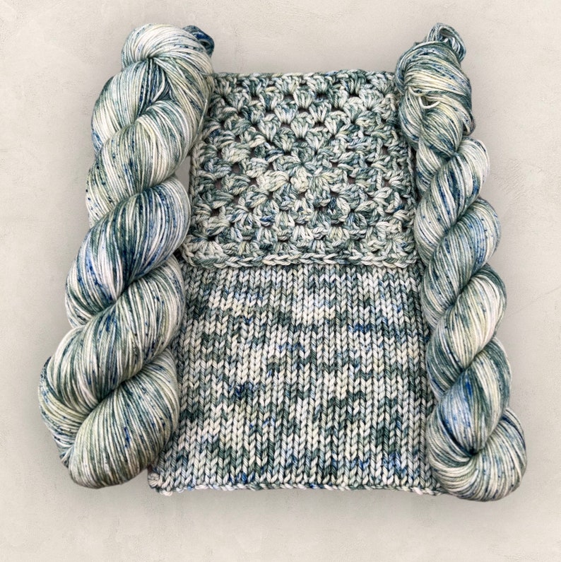 May include: Two skeins of yarn in a green, blue, and white colorway. The yarn is wrapped around a crocheted piece of fabric in a similar colorway.