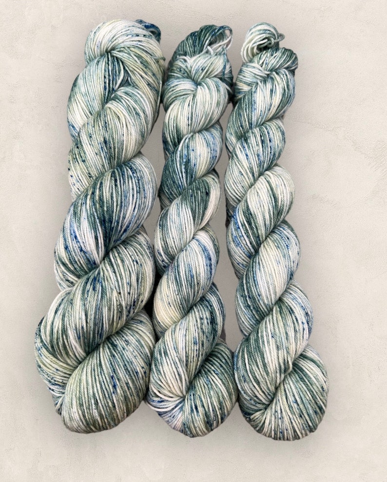 May include: Three skeins of yarn in a green and white speckled pattern. The yarn is wound into a ball shape and is ready to be used for knitting or crocheting.