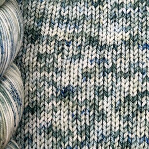 May include: A skein of yarn in a blue, green, and white colorway. The yarn is next to a knitted fabric swatch in the same colorway.