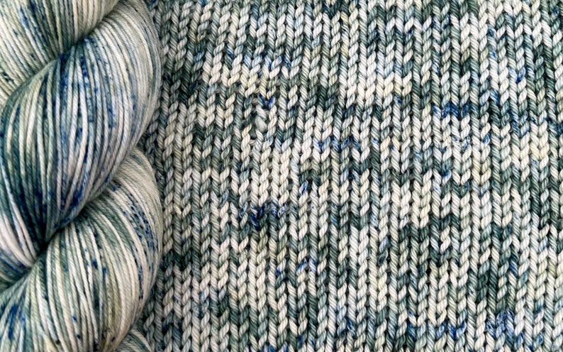 May include: A skein of yarn in a blue, green, and white colorway. The yarn is next to a knitted fabric swatch in the same colorway.