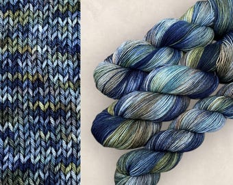 Op de afbeelding: Een streng handgeverfd garen met een gevarieerd kleurenpalet van diep marineblauw, leiblauw en olijfgroen. Het garen wordt getoond in een gedraaide streng naast een staal van tricotsteek breiwerk dat het kleurpatroon weergeeft.