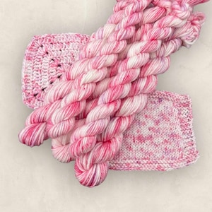May include: A selection of pink and white yarn skeins, a crocheted square, and a knitted square. The yarn is a variegated mix of pink and white. The crocheted square has a geometric pattern. The knitted square has a speckled design.
