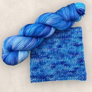 May include: A skein of blue variegated yarn and a knitted swatch. The yarn is a blend of blues and white, with a soft, textured appearance. The knitted swatch displays the same colour variations, showcasing the yarn's potential for knitting projects.