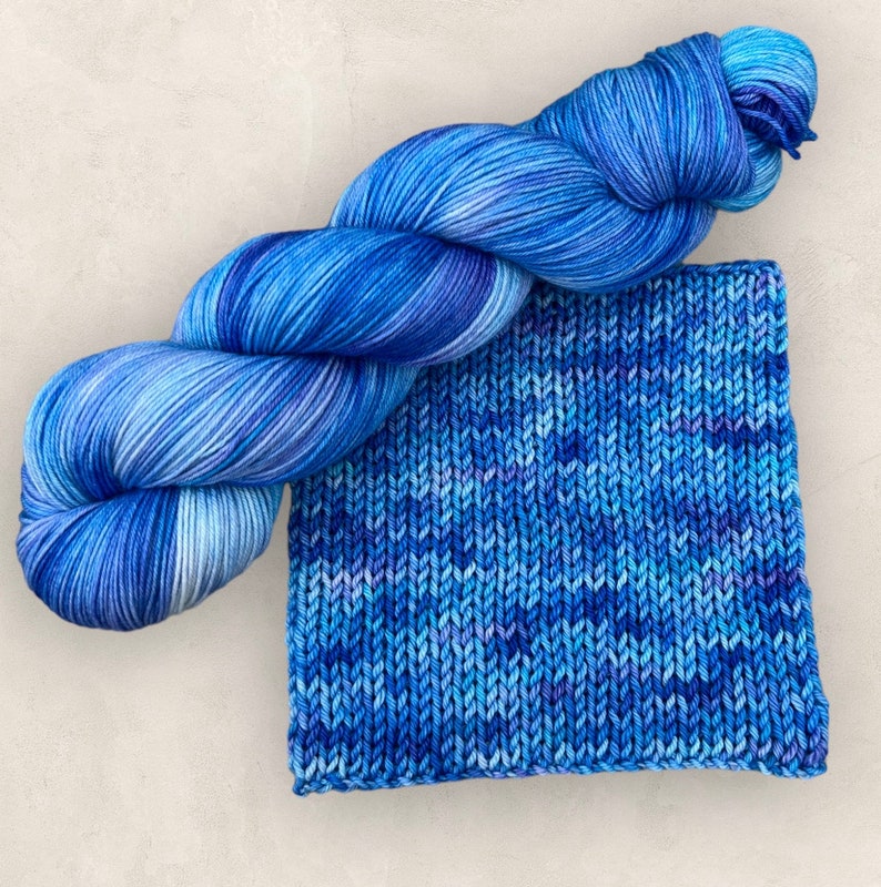 May include: A skein of blue variegated yarn and a knitted swatch. The yarn is a blend of blues and white, with a soft, textured appearance. The knitted swatch displays the same colour variations, showcasing the yarn's potential for knitting projects.